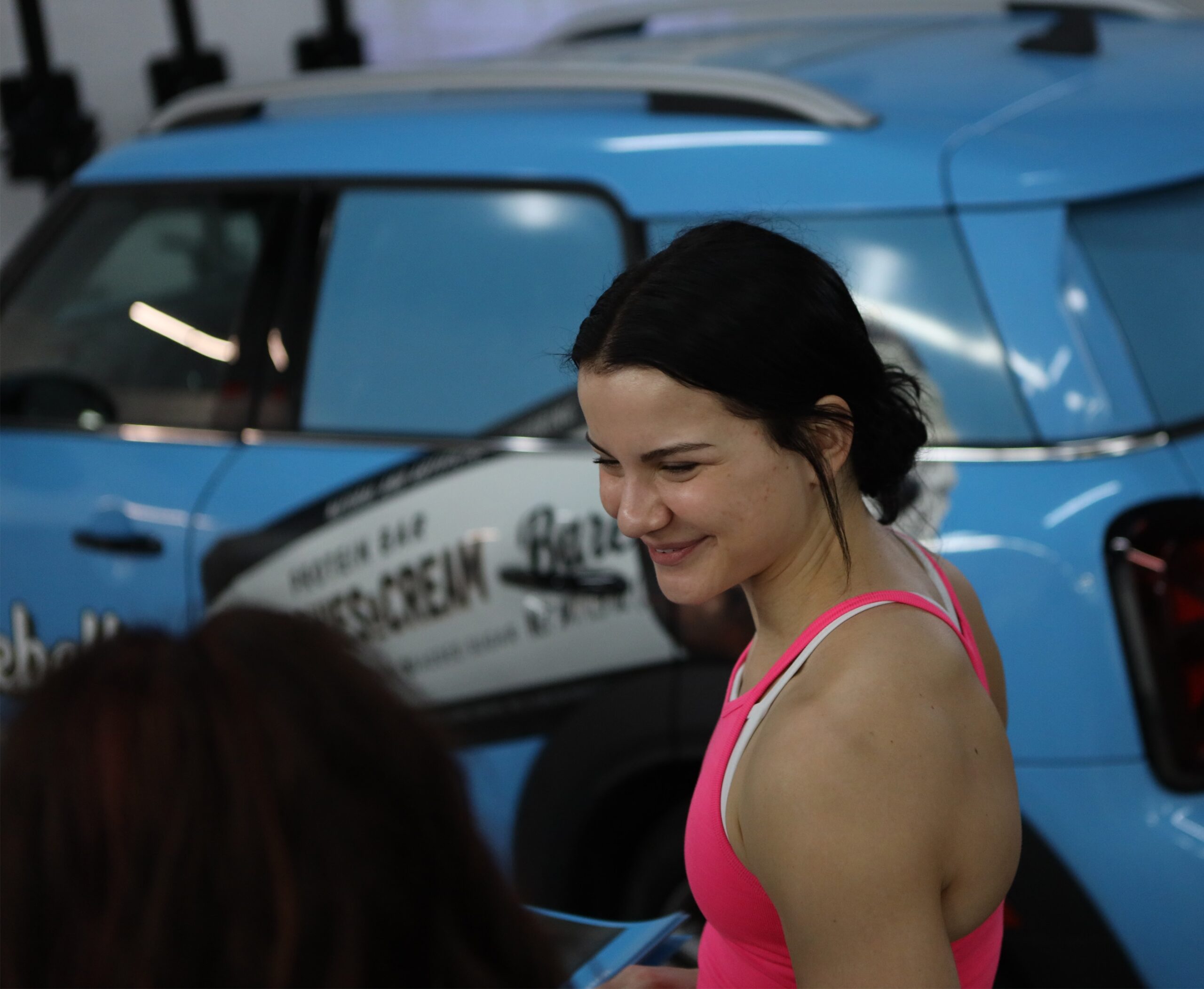 Woman in pink tank top smiling near a blue car.
